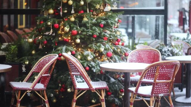 Cozy Parisian cafe terrace with red rattan chairs and round tables covered in fresh snow. A beautifully decorated Christmas tree with lights and ornaments stands in background as snow falls heavily.