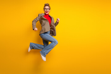 Cheerful young woman jumping in stylish casual wear set against bright yellow background,...