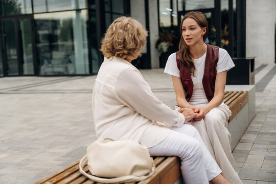 Young woman in casual attire converses with an older woman on a wooden bench outside a modern building with glass facade and urban surroundings