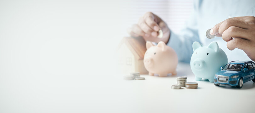 Person dropping coins into piggy banks with house and car models on desk, concept of financial planning, saving money for home, vehicle purchase, investment, and future security.