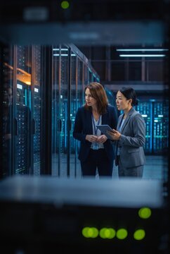 Two businesswomen reviewing a tablet beside server racks, framed through equipment