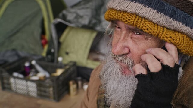 Senior Caucasian homeless man answering smartphone outdoors near tent shelter. Footage suitable for poverty awareness, charity campaigns, social welfare, non-profit initiatives