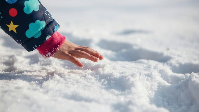 A child in a colorful winter jacket gently touches fresh snow outdoors in bright daylight. Perfect for winter holidays, childhood memories, and seasonal greeting card designs.