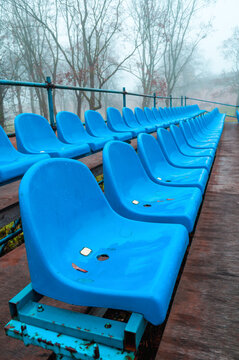 Autumn stadium background. Blue plastic seats arranged in rows on a wooden platform. Bare trees in thick fog behind the rusty metal frame structure