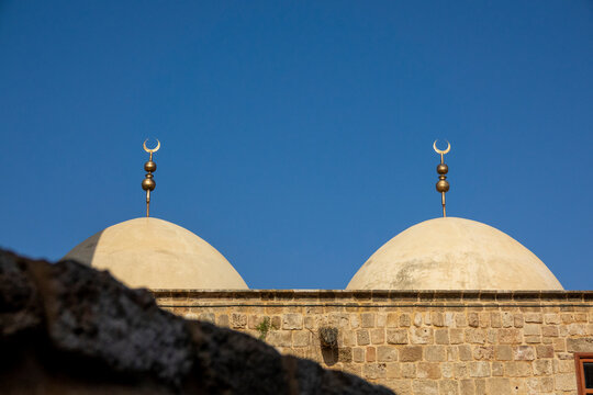 Twin mosque domes topped with golden crescent finials rise above an old stone wall against a clear sky, South Governorate, Tyre, Lebanon