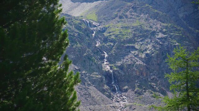 Serene mountain valley with a cascading waterfall. Tall coniferous trees frame the scene. Rocky slopes rise steeply behind the water flow. Sunlight illuminates parts of the canyon