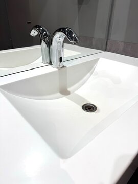 Modern automatic touchless faucet on white ceramic sink in public restroom. Sensor tap for hand washing and hygiene. Smart bathroom technology and water saving concept.