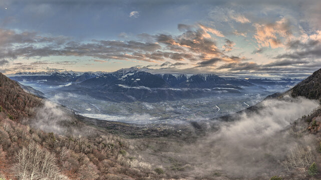 Aerial view of Valtellina valley with mist rolling over the landscape, snow-capped mountains in the distance, and sunset light over the town of Sondrio, Lombardy, Italy.
