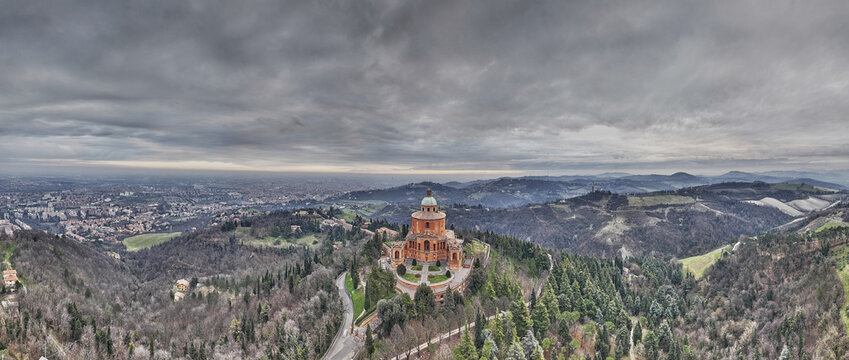 Aerial view of the Sanctuary of the Madonna of San Luca on a hill overlooking the city, surrounded by forests and hills under a dramatic cloudy sky, Bologna, Emilia-Romagna, Italy.