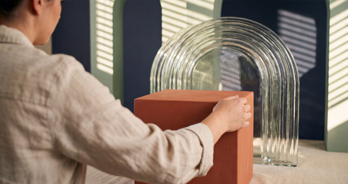 Woman prop stylist arranging a terracotta display block in a photography studio with modern glass arches and sunlight shadows