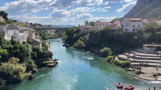 Scenic high angle view of the emerald green Neretva River in Mostar, Bosnia and Herzegovina, capturing a person jumping off a training diving platform into the river near the historic Old Bridge area.