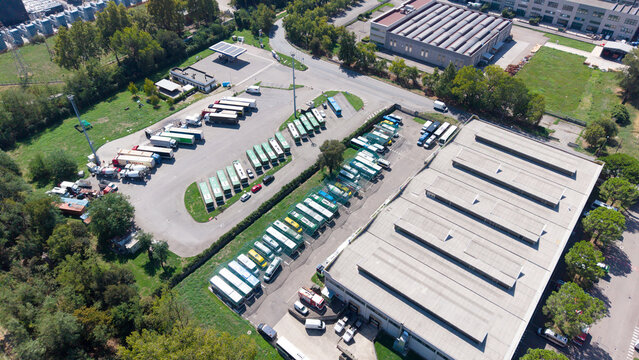 Aerial view of a fleet of buses and heavy machinery parked in a depot with industrial buildings and surrounding greenery in Milan, Lombardy, Italy.