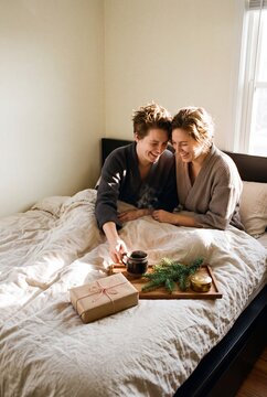 Happy Lesbian Couple Laughing in Bed with Morning Coffee and Christmas Gift During Cozy Winter Holiday