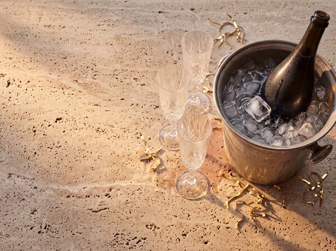 Bottle of champagne chilling in a metal ice bucket with crystal glasses and gold confetti on a textured stone table