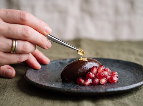 Pastry chef using tweezers to decorate a dark chocolate quenelle with edible gold leaf and fresh pomegranate seeds on a rustic plate