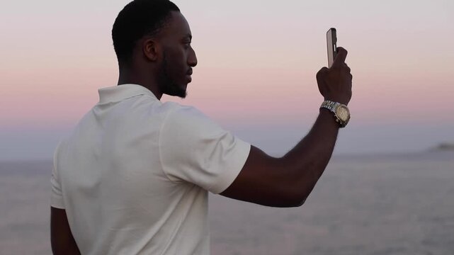 Man recording coastal landscape with smartphone at dusk. Man standing by the sea at dusk, slowly recording the surrounding coastal landscape with a smartphone.