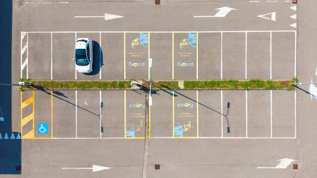 Aerial view of an electric vehicle charging station with a white car parked in a designated spot, featuring painted markings and directional arrows in Milan, Lombardy, Italy.