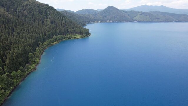 Aerial view of Lake Tazawa featuring deep blue water surrounded by lush green forested mountains in Semboku, Akita, Japan.