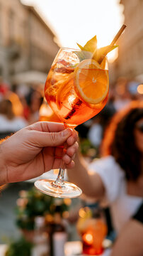 Hand holds a glowing orange aperitivo spritz aloft at golden hour, blurred piazza backdrop capturing warm social evening celebration atmosphere.