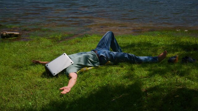 Person lies on grass by water with laptop over face. Person appears unaware or relaxed. Scene suggests digital detox or work break. Ideal for themes of relaxation, technology break, or nature escape