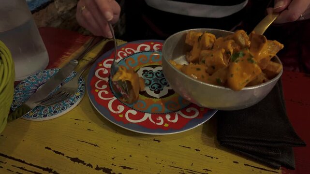 A person serves warm patatas bravas from a metal pan onto a colorful plate. Authentic traditional tapas celebrate the joy of sharing delicious culinary experiences in a cozy social environment.
