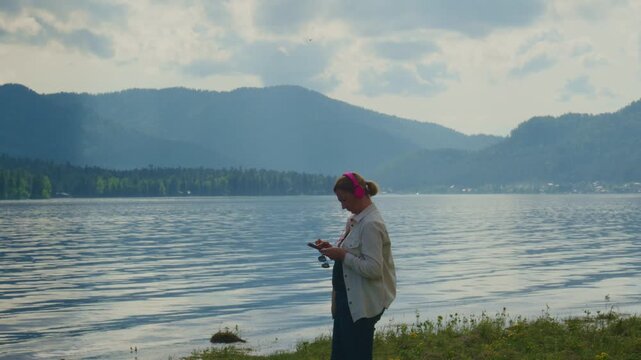 Woman stands near a calm lake, wearing a red headscarf and light jacket. She looks at her phone, seemingly relaxed and thoughtful. Behind her, gentle waves ripple against the shore