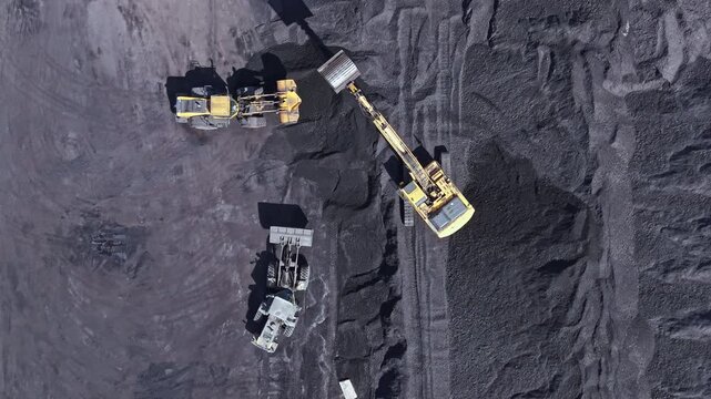 Three large machines work on a coal mining site, moving and loading coal. The setting shows dark ground covered in coal. The sun shines down on the busy site.
