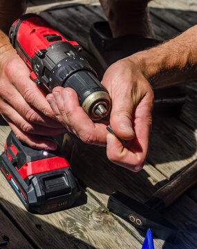 Close-up of hands changing a screwdriver bit in a cordless drill chuck, with battery pack resting nearby on wooden surface outdoors . High quality photo