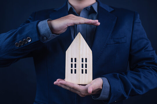 Business professional protecting a wooden house model with his hands, symbolizing home insurance, property protection, security, mortgage, real estate, investment, ownership, safety, and financial pla