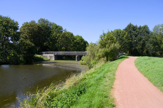 Red bicycle path running along a river bank towards a small weir bridge in a green countryside landscape under a clear blue sky.
