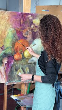 Teenage girl in college painting class, art studio, art workshop. She paints still life on canvas with oil paints, holds brush.