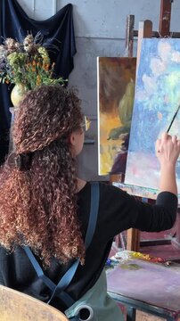 Teenage girl in college painting class, art studio, art workshop. She paints still life on canvas with oil paints, holds brush.