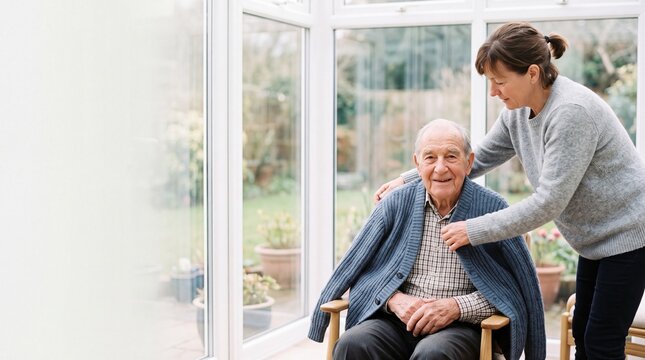 Caring adult daughter helping smiling senior father put on a warm cardigan in sunroom at home