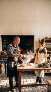 Professional senior baker sifting flour over artisan bread loaves alongside his young apprentice in front of a traditional wood-fired oven