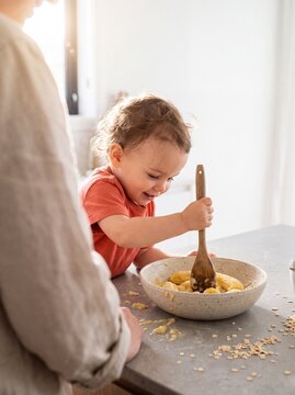 Happy toddler mashing bananas in a bowl with a wooden spoon while cooking healthy breakfast in a bright kitchen with mother