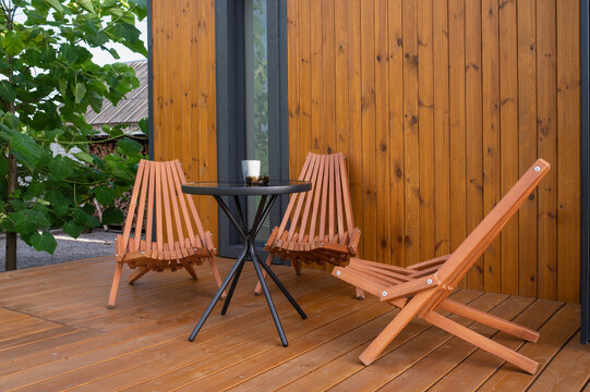 Folding lounge chairs and a glass table on a wooden deck; a backyard garden relaxation area in summer.