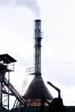 An old metal industrial factory chimney stack emitting white smoke into the sky, representing manufacturing activity and air pollution issues.