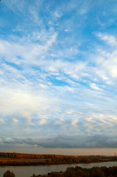Nature landscape background. A wide river with autumn trees on the banks. Layered grey and white clouds above under a blue sky in a vertical view