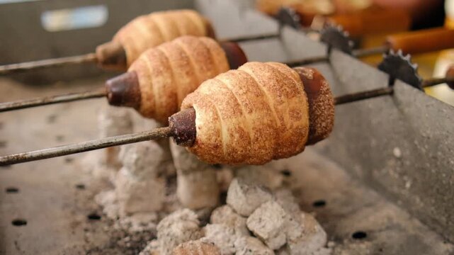 Sweet pastries on skewers roast over hot coals in a street food environment, highlighting their golden-brown layers and crispy texture, trdelnik