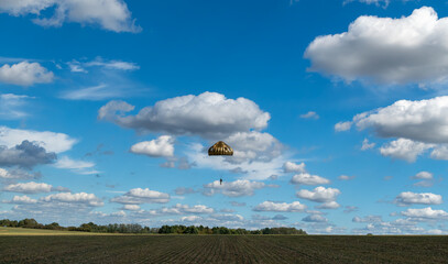 A German Bundeswehr paratrooper descending over a plowed field under a round parachute against a wide blue sky with fluffy white clouds. © Frank
