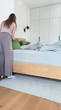 Making bed in a minimalist bedroom during morning housekeeping routine. Young female householder arranges pillows and adjusts blankets on a contemporary wooden frame. Clean home environment