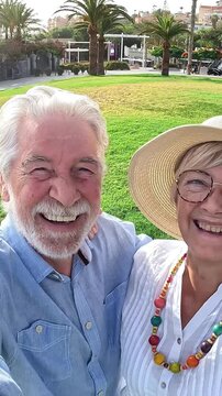 Cheerful elderly couple having fun together taking a selfie video in a beautiful public park with palm trees, enjoying their retirement and creating lovely memories of an outdoor summer vacation