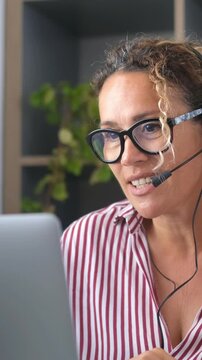 Smiling businesswoman wearing a headset with a microphone working on a laptop, providing customer service from her home office, confidently handling client inquiries and support