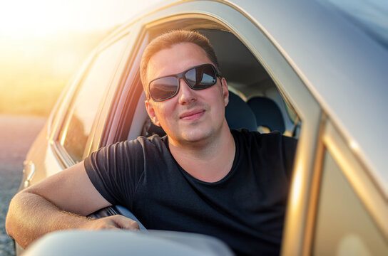 A happy man wearing sunglasses looks out the window of his car during a sunset road trip.