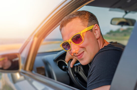 A happy man wearing yellow sunglasses looks out the open window of his car during a sunny road trip.
