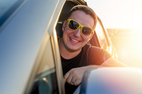 A cheerful man wearing yellow sunglasses smiles out the open window of a car during a sunny day road trip.