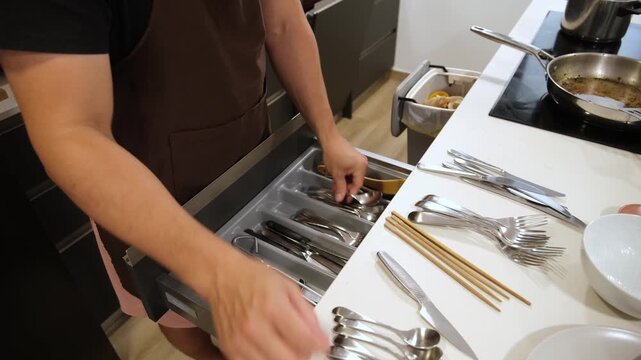 Man wearing an apron tidies up and sorts clean cutlery into a kitchen drawer organizer