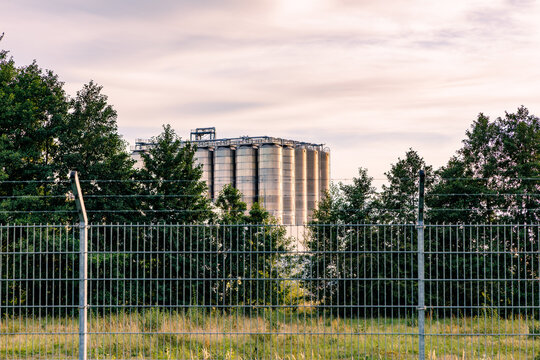 Industrial storage silos and tanks seen behind metal security fence surrounded by green trees at dusk, factory security and energy storage concept