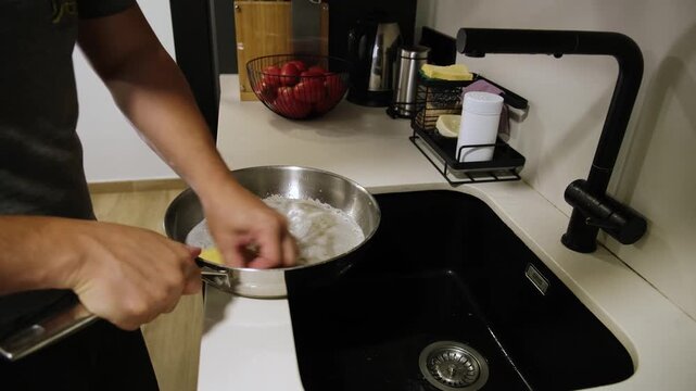 Male hands washing a stainless steel frying pan with a sponge in a modern black kitchen sink