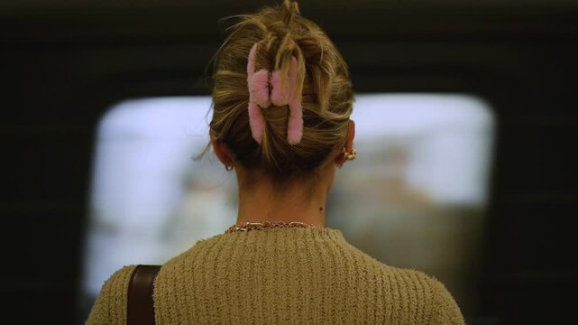 A woman in a sweater stands at a subway station, facing forward with a pink hair clip holding her hair. She appears to be waiting for a train as motion blur suggests nearby movement